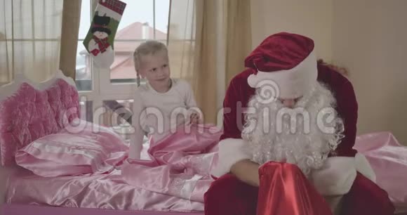 穿着圣诞老人服装的白人男子坐在粉红色的床上小女孩扎着辫子抚摸着他的肩膀视频的预览图