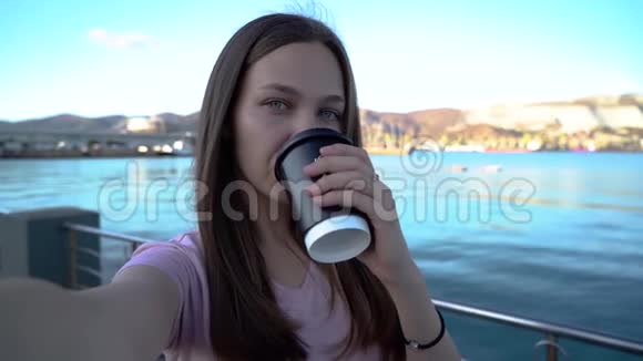 迷人的年轻女子穿着粉红色的t恤喝着咖啡用手机在海滩上带着山脉和山脉视频的预览图