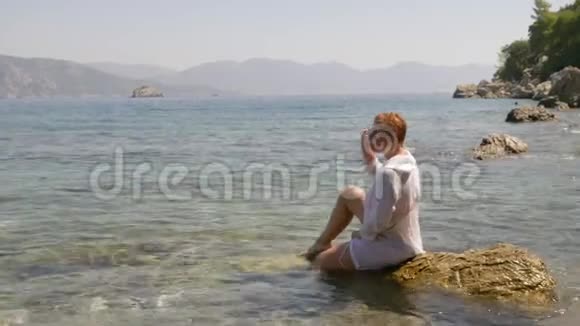 夏季旅行时坐在大海中的大石头上的放松的女人在山景上挥手快乐的女人享受柔软的海洋视频的预览图