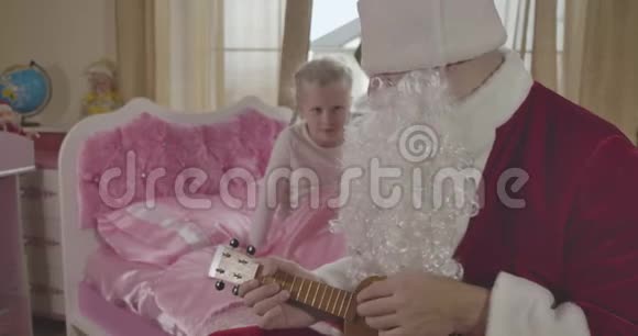 可爱的高加索儿童坐在粉红色的床上看着圣诞老人玩四弦琴穿圣诞老人服装的成年男子视频的预览图