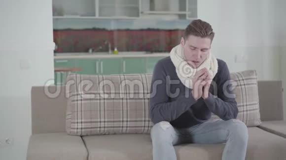 白人男子戴着白色围巾坐在沙发上打喷嚏的肖像年轻女子端着一杯热饮料来疾病视频的预览图