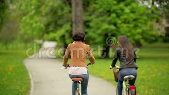 穿着夹克和牛仔裤戴深色头发的积极微笑的女性朋友骑着自行车两个漂亮的背面视频的预览图