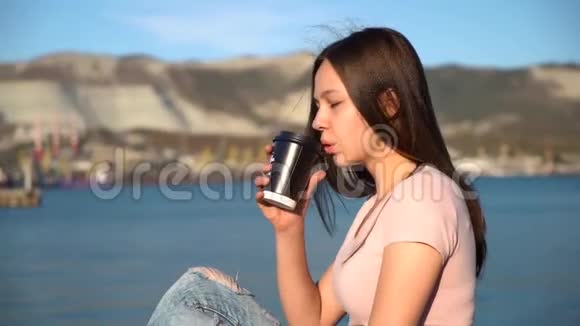 漂亮的女孩穿着粉红色的t恤坐在河堤上的栏杆上喝着黑杯子里的咖啡视频的预览图