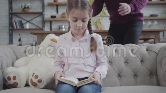 可爱的白种人女孩坐在沙发上带着泰迪熊和看书父亲过来闭上眼睛的肖像视频的预览图