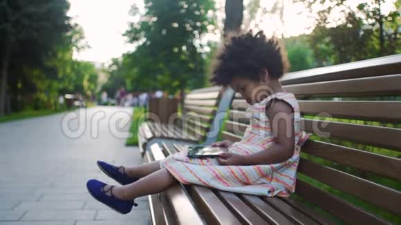 小非洲裔美国女孩正坐在公园的长凳上在平板电脑上看动画片视频的预览图