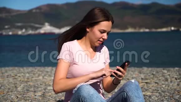 快乐迷人的女孩穿着粉红色的t恤和蓝色牛仔裤或者女人坐在海滩上用智能手机聊天慢动作视频的预览图
