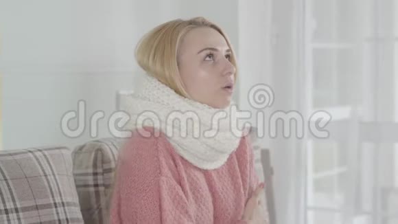 年轻迷人的白人妇女戴着白色围巾颤抖的肖像金发女孩坐在沙发上打喷嚏视频的预览图