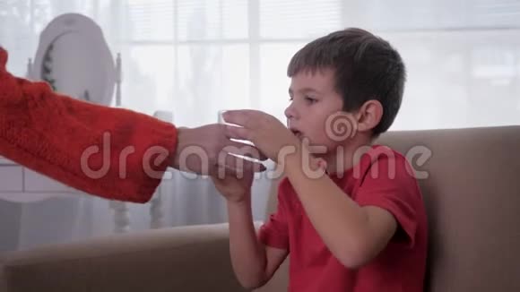口渴的小男孩在家里坐在沙发上从妈妈手里拿一杯水喝视频的预览图