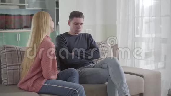 积极的高加索女人触摸丈夫和微笑坐在沙发上的男人被他的配偶激怒了关系视频的预览图