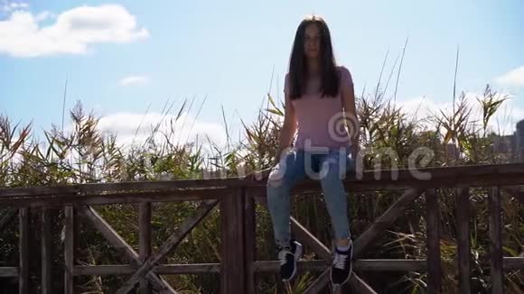 漂亮漂亮的女孩穿着粉红色的t恤和撕破的蓝色牛仔裤坐在栏杆上整理头发视频的预览图