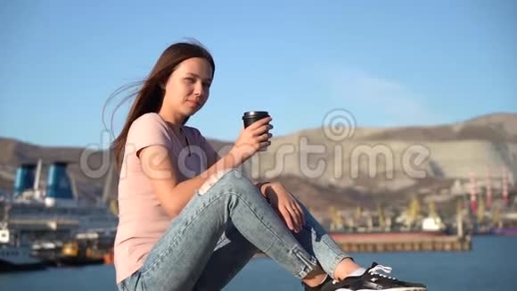 漂亮的女孩穿着粉红色的t恤坐在河堤上的栏杆上喝着黑杯子里的咖啡视频的预览图