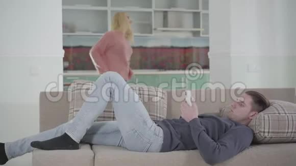年轻的高加索男人躺在沙发上用智能手机的肖像而他不幸的妻子在后台抽真空视频的预览图
