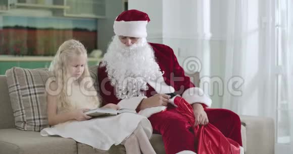 漂亮的金发白种人女孩穿着优雅的白色连衣裙和皇冠和圣诞老人坐在马车上看书圣诞老人视频的预览图