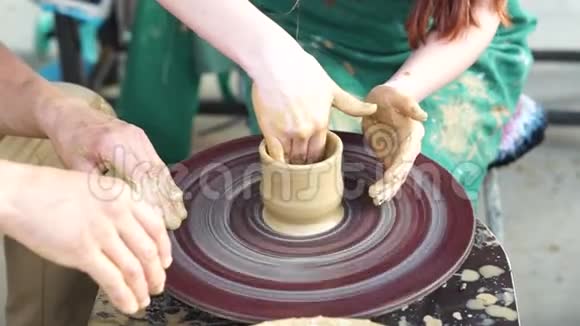 主人晒黑的手和一个围着绿色围裙坐在陶工轮上的女孩的手做成了一壶粘土视频的预览图