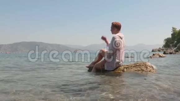 一个穿着白色湿衣的女人坐在海浪中的石头上快乐的女游客坐在海边的岩石上视频的预览图