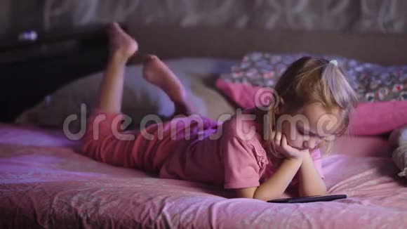 可爱的金发小女孩穿着粉红色睡衣躺在床上用智能手机看动画片幼儿女孩子视频的预览图
