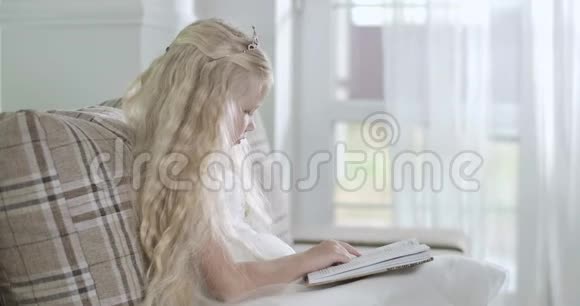 侧面观看金发白种人小女孩坐在沙发上看书圣诞老人来到玻璃门敲门视频的预览图