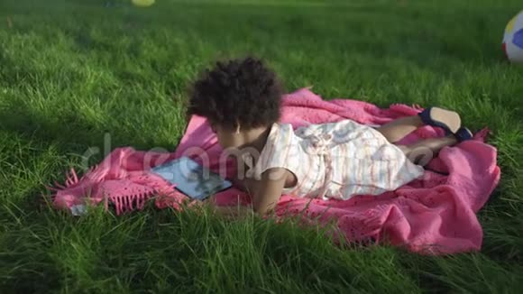 一个非洲裔小女孩躺在公园里躺在床上在平板电脑上看动画片视频的预览图