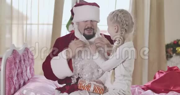 可爱的白种人女孩扎着辫子手里拿着假胡子就像穿着圣诞老人服装的男人抱着她的膝盖一样视频的预览图