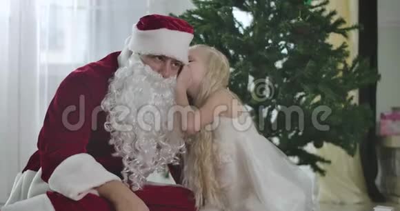 漂亮的白人女孩和圣诞老人坐在家里的圣诞树前在他耳边低语儿童问题视频的预览图