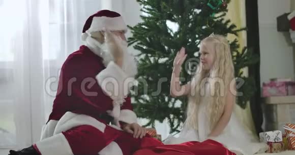 可爱的白种人女孩穿着白色连衣裙戴着皇冠和圣诞老人坐在新年树前给他击掌视频的预览图
