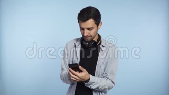年轻自信的男人拿着手机和滚动屏幕或打字信息隔离在蓝色背景上戴着耳机视频的预览图