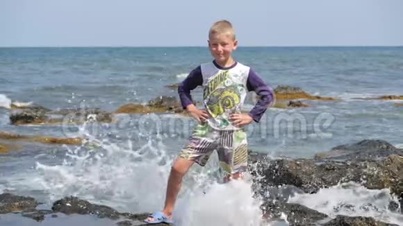 男孩站在被海浪的浪花冲走的岩石上波浪在岸边折断拇指向上举起婴儿视频的预览图