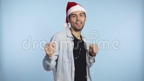 戴着圣诞红帽子戴着花环的英俊男子正对着镜头蓝色的墙壁背景跳舞微笑视频的预览图