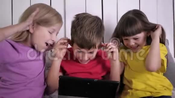 快乐的孩子们穿着多色的衣服在平板电脑前坐在床上在家里放松视频的预览图