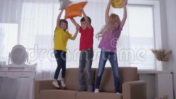 活跃的孩子们玩得很开心他们在沙发上跳着手里拿着枕头在灯光下靠着窗户视频的预览图