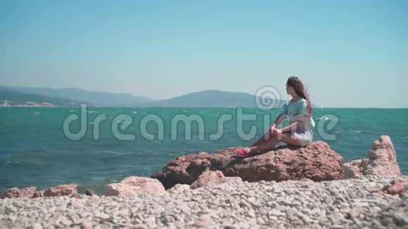 一个穿着蓝色T恤和灰色工作服的女孩坐在海边的石头上视频的预览图