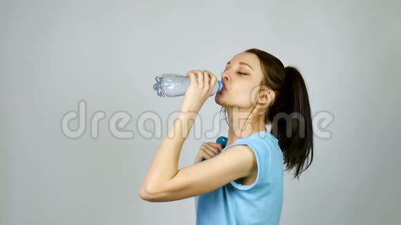 一个长着深色头发的布鲁内特女孩在灰色的环境下锻炼后正在用塑料透明瓶子喝纯净水视频的预览图