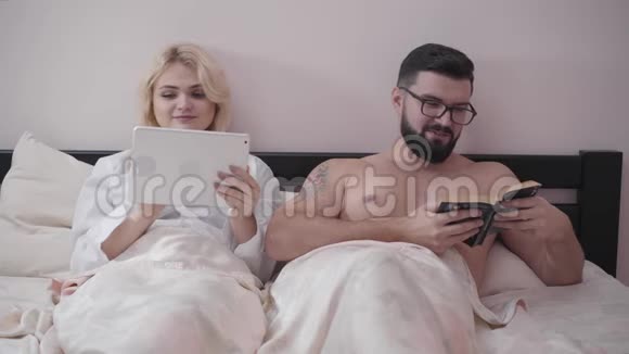 幸福的白种人配偶晚上躺在床上的肖像可爱的金发女人用平板电脑男人看书妻子视频的预览图