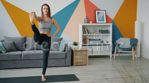穿着紧身裤和单腿平衡的漂亮女人在家练习瑜伽视频的预览图