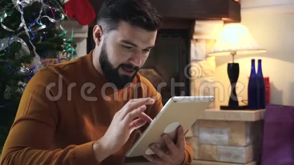 集中白人男子的肖像黑胡子坐在圣诞树前使用平板电脑严重视频的预览图