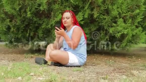 一个扎着红辫子的年轻女人正坐在草地上的公园里看着她的手机移动摄像机视频的预览图