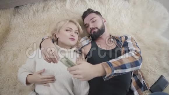 快乐的富加索夫妇躺在柔软的地毯上数着美元穿着白毛衣的金发女孩和英俊的胡子视频的预览图