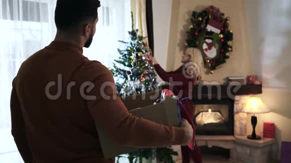 镜头跟随成年白人男子带着盒子走到美丽的女人装饰圣诞树丈夫带来视频的预览图