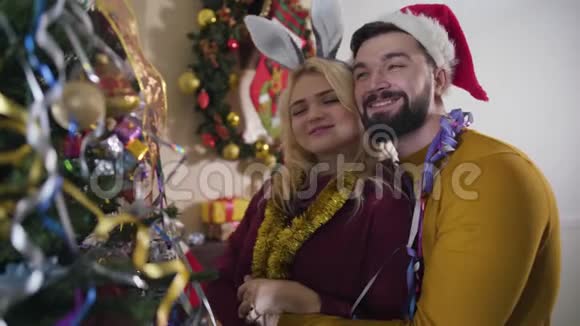 快乐的白人年轻夫妇拥抱在圣诞树前微笑英俊的男人和金发女人视频的预览图
