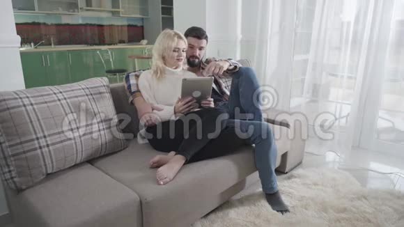 阳性白种人男女坐在沙发上使用平板电脑丈夫和妻子在网上购买视频的预览图