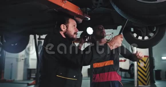 一个成熟的机械师和他的助手在车库里穿制服在损坏的汽车下试图找到问题视频的预览图