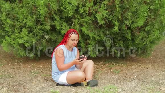 一个扎着红辫子的年轻女子坐在草地上的公园里看着她的手机视频的预览图