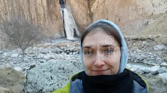 在凉爽的天气里一个穿着连帽衫的女孩在高加索瀑布的背景下自拍冷冻女人旅行视频的预览图