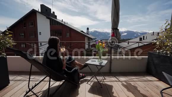 年轻女子在阳台上喝果汁在背景上喝雪山假期和放松视频的预览图