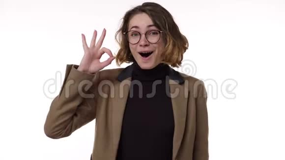 穿着棕色夹克的漂亮年轻女子微笑着竖起大拇指在白色背景上眨着眼睛视频的预览图