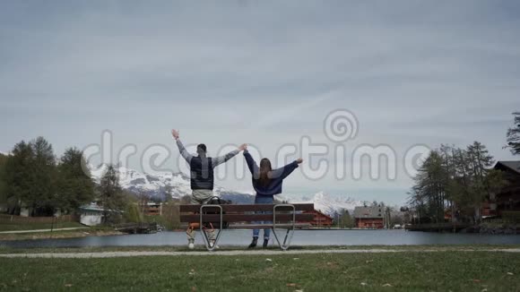 相爱的幸福夫妇站在一起双手举起在湖边的木凳上美丽的山景后方视频的预览图