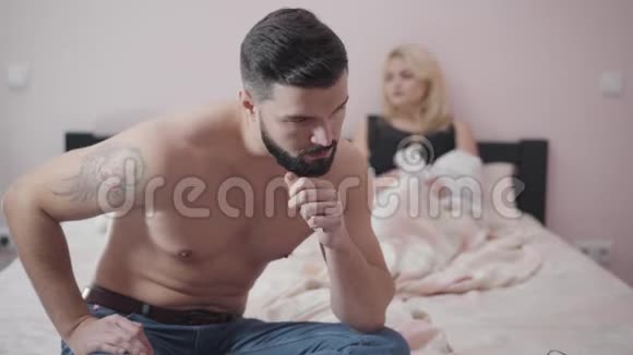 恶心的白种人男人坐在床上回头看妻子或女朋友悲伤模糊的女人躺在后台视频的预览图