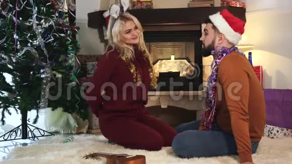 积极的金发白种人女人和丈夫坐在壁炉和圣诞树前背着礼物视频的预览图