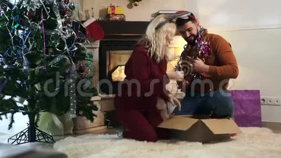开朗的白种人金发女人和长胡子的男人坐在壁炉和圣诞树前玩四弦琴视频的预览图