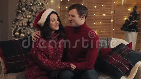 喜笑颜开的情侣坐在靠近圣诞树的客厅的沙发上看镜头视频的预览图